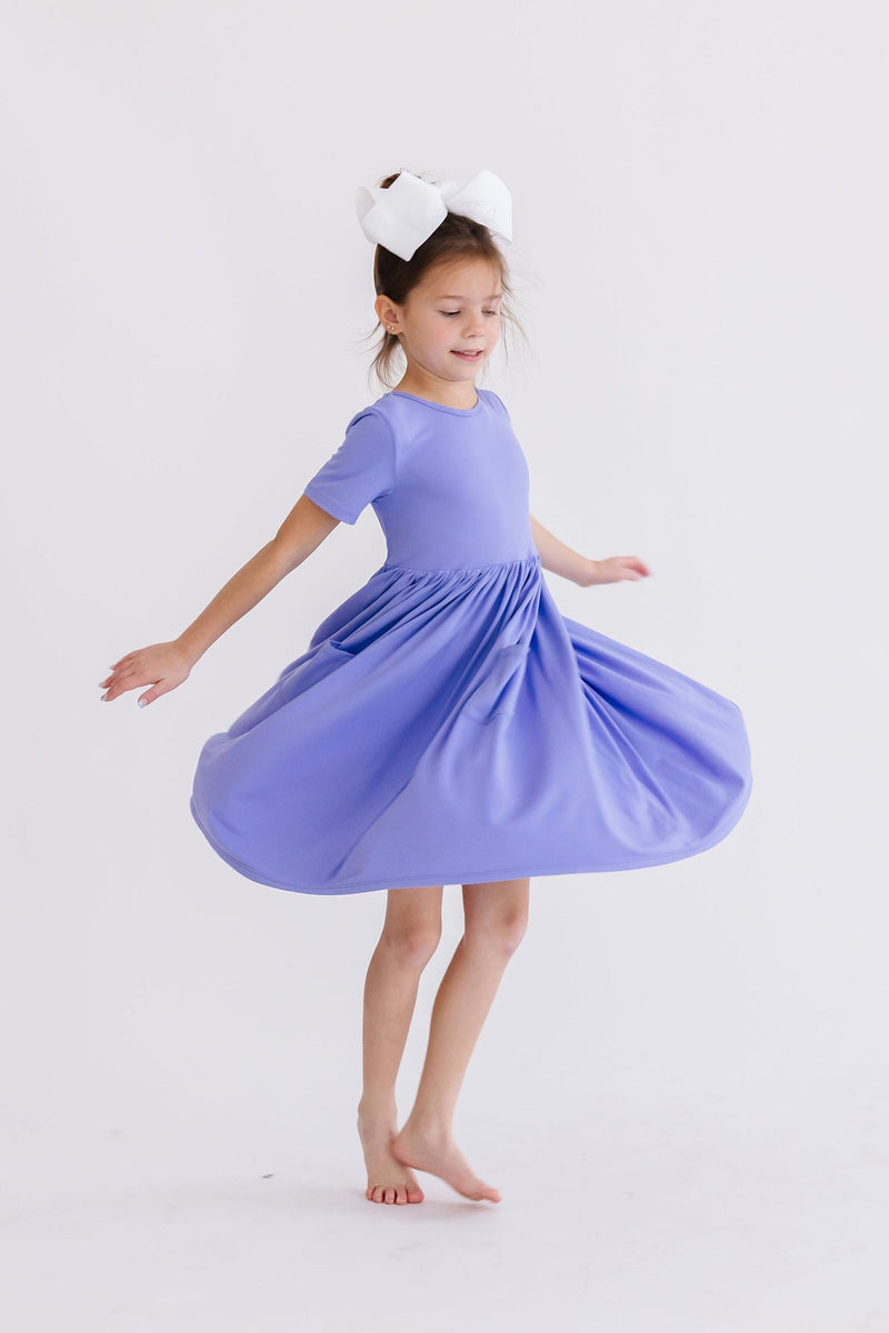 Delighted child in a Periwinkle S/S Pocket Twirl Dress, spinning around to showcase the dress's twirl-ability, ideal for joyful playtime.