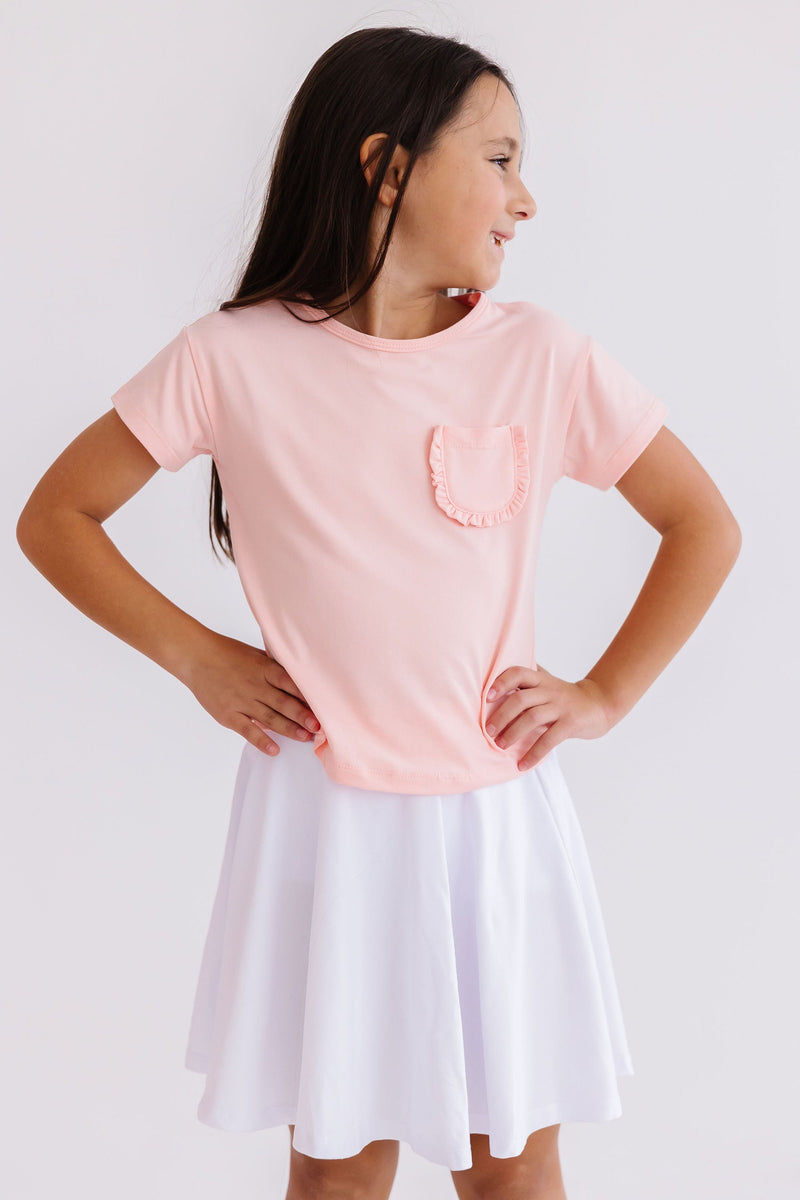 Happy girl posing in the Peach Ruffle Pocket Tee, perfectly paired with a flouncy white skirt for a chic look.