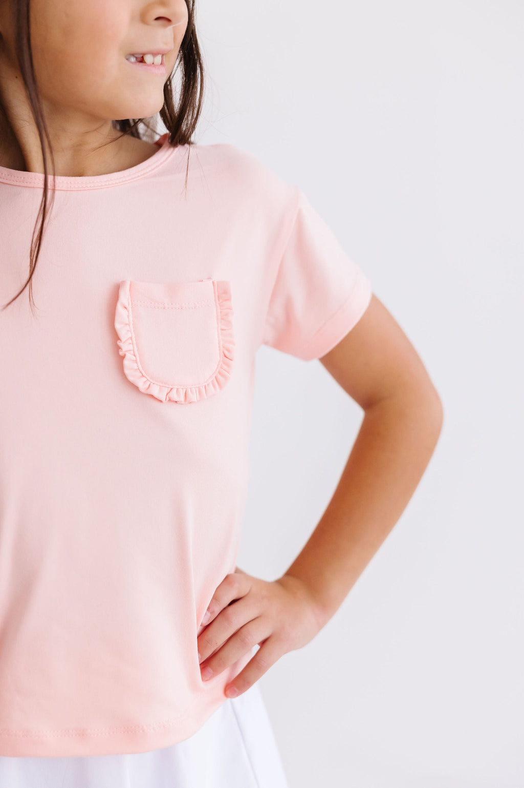 Charming young girl showcasing the Peach Ruffle Pocket Tee with playful ruffled pocket detail, exuding stylish confidence.