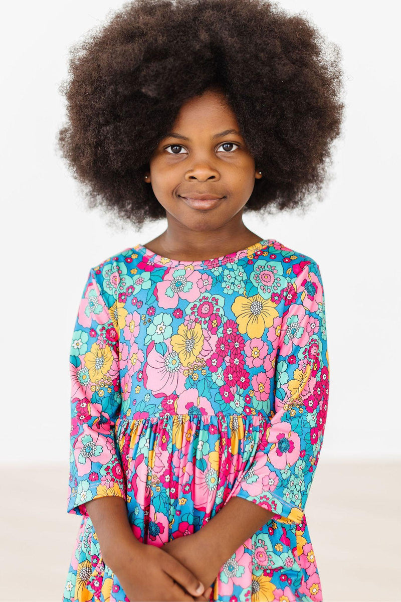 A young girl poses confidently in the Feeling Floral 3/4 Sleeve Pocket Twirl Dress, showcasing a vibrant floral design and an adorable smile.
