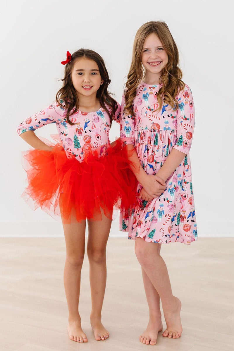 Two girls showcasing the Sugarplum Fairy 3/4 Sleeve Pocket Twirl Dress; one wears a vibrant red tulle skirt, while the other models the dress with a fun festive print.