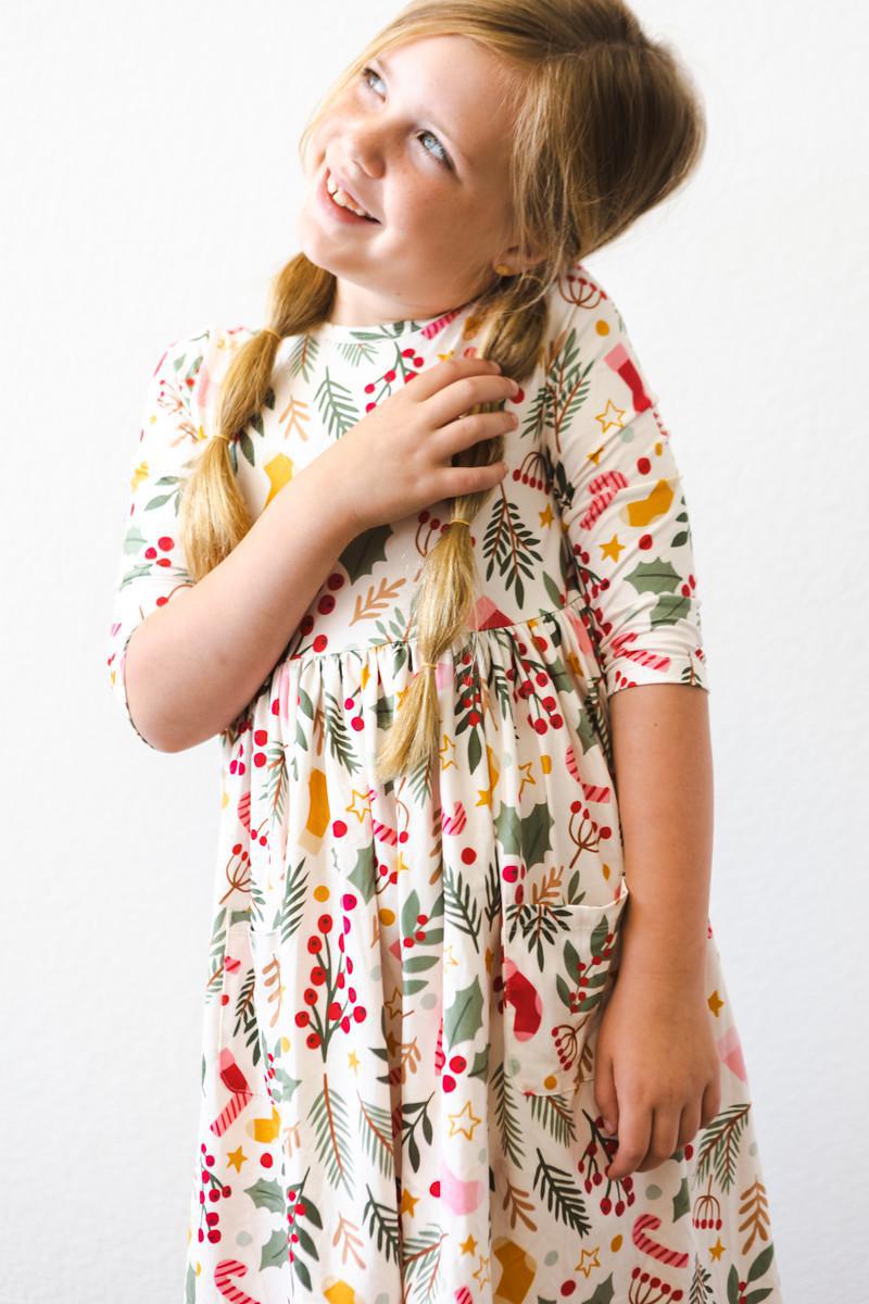 A cheerful young girl in the 'Holly Jolly Christmas' 3/4 Sleeve Pocket Christmas Twirl Dress for Girls, looking up with a bright smile.