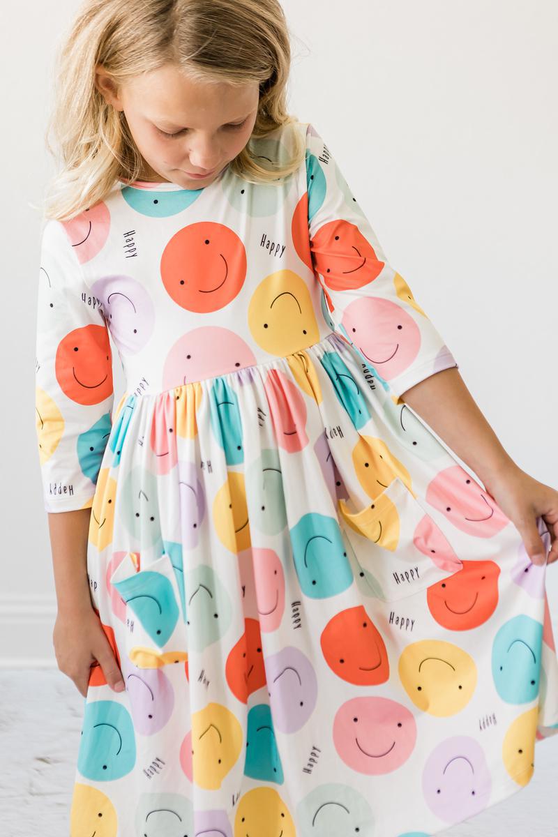 A smiling girl wearing the 'Happy Days' 3/4 Pocket Fun Twirl Dress for Girls, featuring colorful smiley graphics, ideal for joyful occasions.