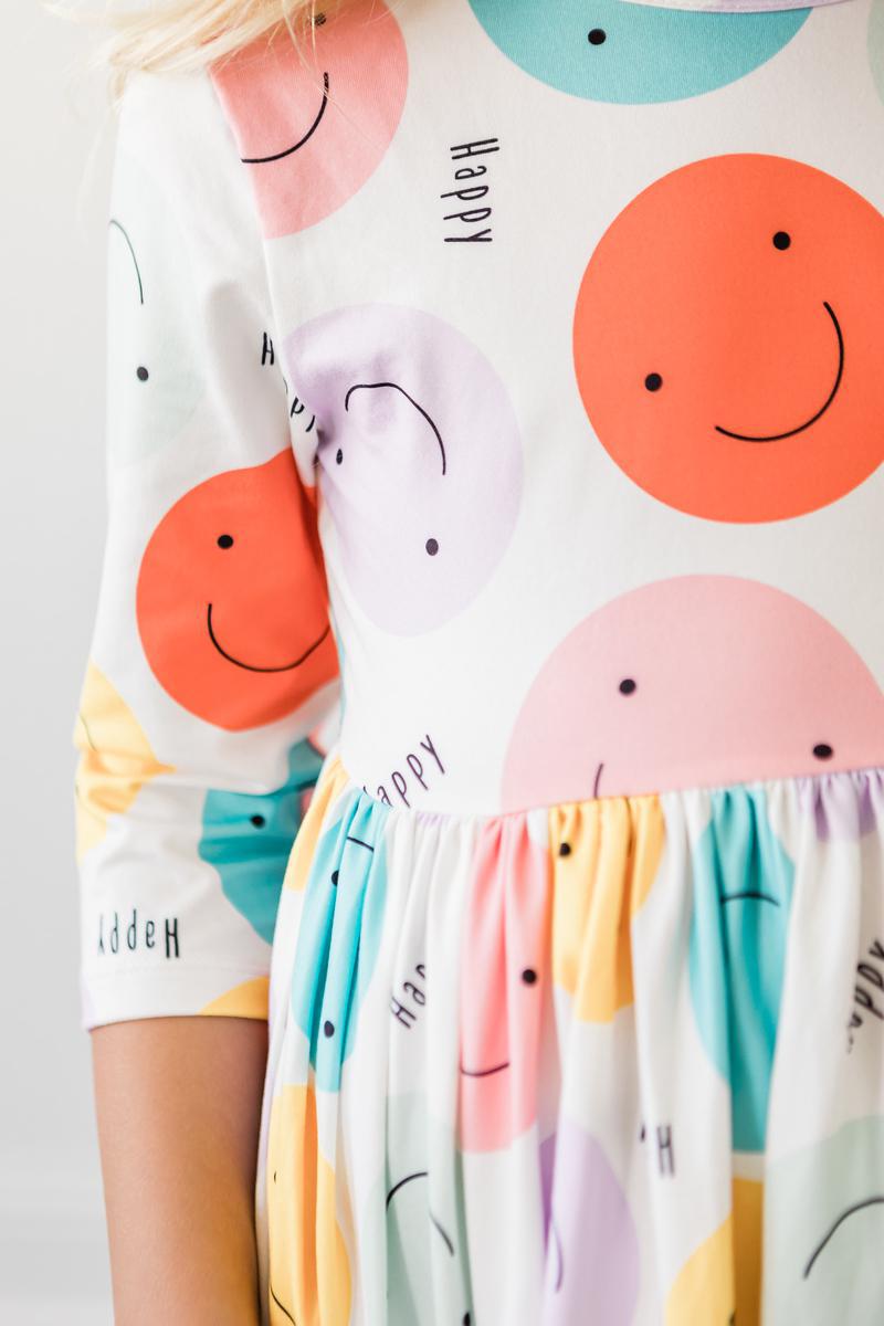 A girl wearing the 'Happy Days' 3/4 Pocket Fun Twirl Dress for Girls, focusing on the upper design with smiley faces and vibrant colors.