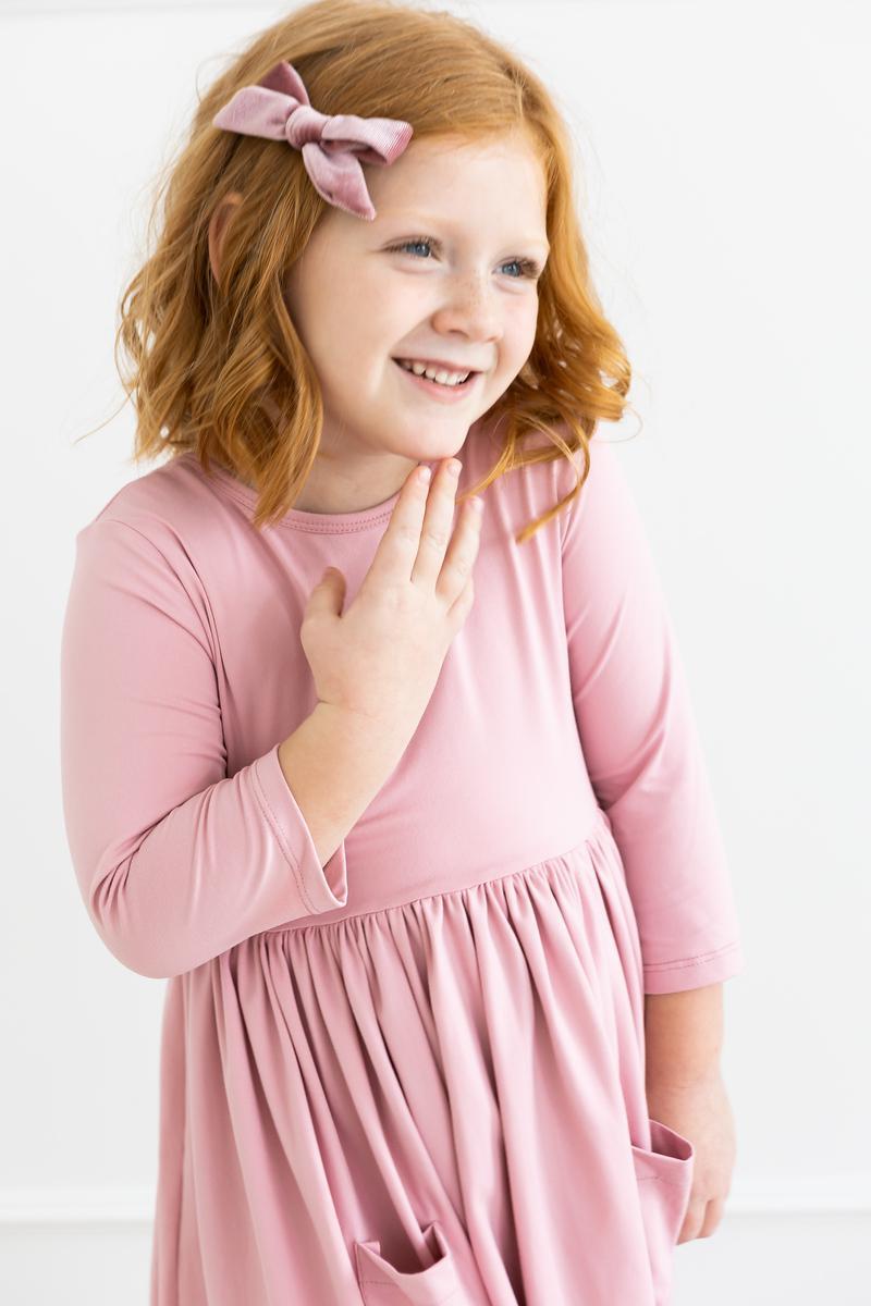 A cheerful girl in the Vintage Pink 3/4 Sleeve Pocket Twirl Dress from Mila & Rose, smiling with a bow in her hair, perfect for any occasion.