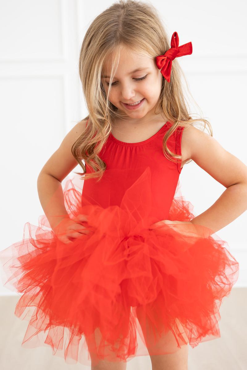 A young girl wearing the Red Tank Tutu Leotard, smiling and standing proudly with hands on her hips, showcasing the vibrant red tutu.