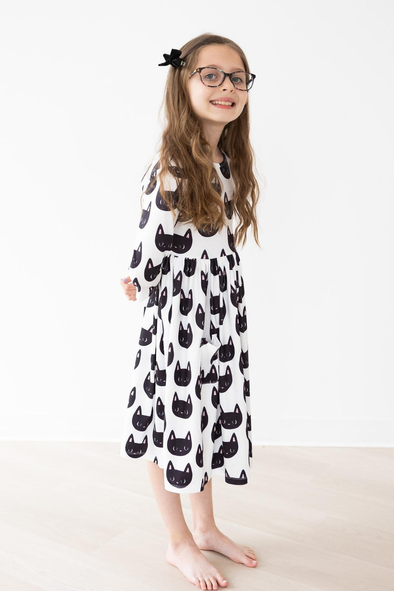 A smiling girl wearing the Purrfect 3/4 Sleeve Pocket Twirl Dress by Mila & Rose, with a lovely black cat print, styled with glasses and a hair accessory.