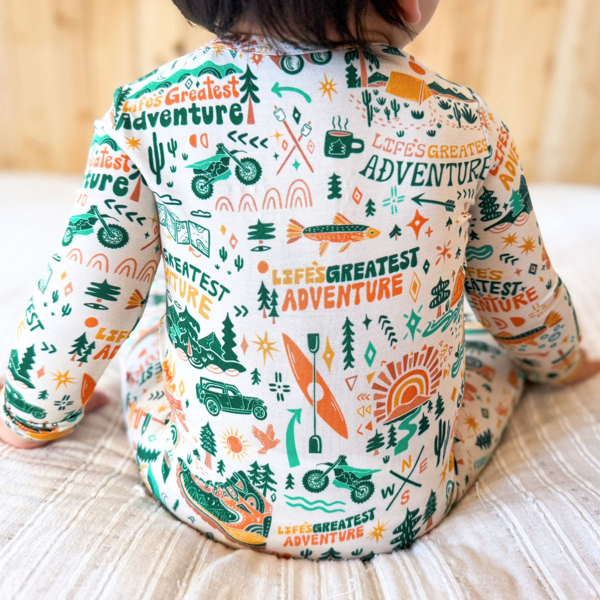Child wearing a long-sleeve shirt with 'Life's Greatest Adventure' print on a wooden floor.