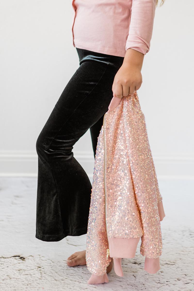 A little girl in Black Velvet Bells by Mila & Rose® holding a sparkling pink jacket, emphasizing the versatility and style of her outfit.