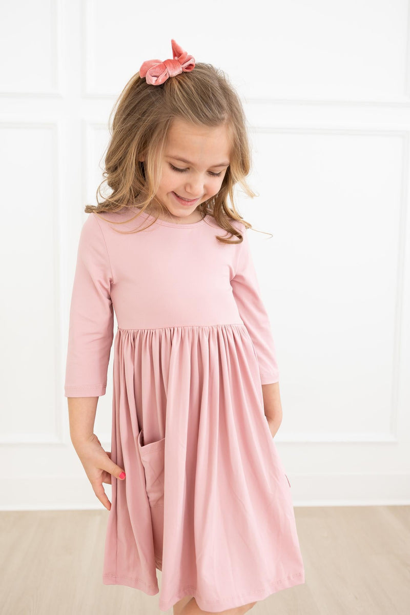 A young girl delightfully examining her Vintage Pink 3/4 Sleeve Pocket Twirl Dress by Mila & Rose®, featuring a relaxed fit and convenient pockets, ideal for fun and style.