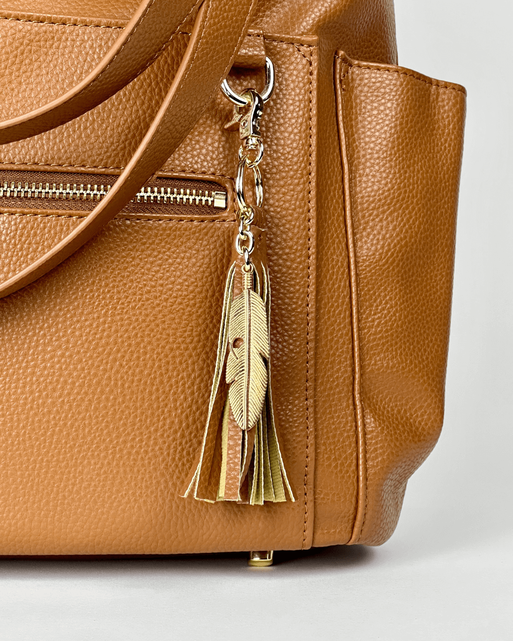 Close-up of cognac pebbled vegan leather side pocket with gold zipper and decorative metallic gold feather tassel charm