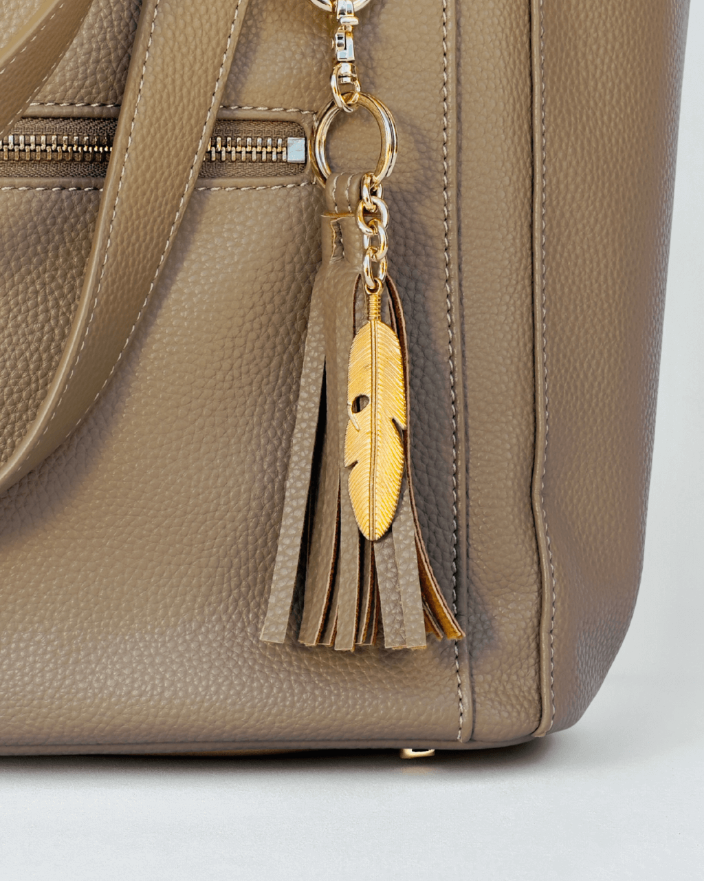 Close-up of taupe pebbled vegan leather texture with gold zipper and engraved leaf tassel detail