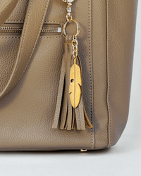 Close-up of taupe pebbled vegan leather texture with gold zipper and engraved leaf tassel detail