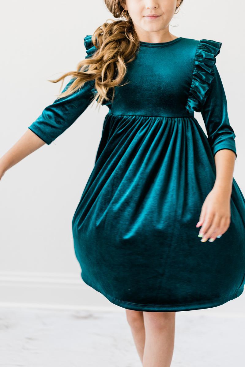 A child wearing the Teal Velvet Ruffle Twirl Dress by Mila & Rose, showcasing its elegant design and soft fabric. The dress features ruffles on the shoulders and a full skirt that enhances its playful look.