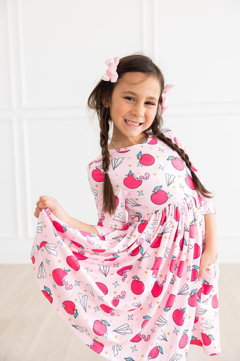 Girl playfully twirling in the Star Student 3/4 Sleeve Pocket Twirl Dress, featuring a cheerful apple design, showcasing fun and elegance in everyday wear.