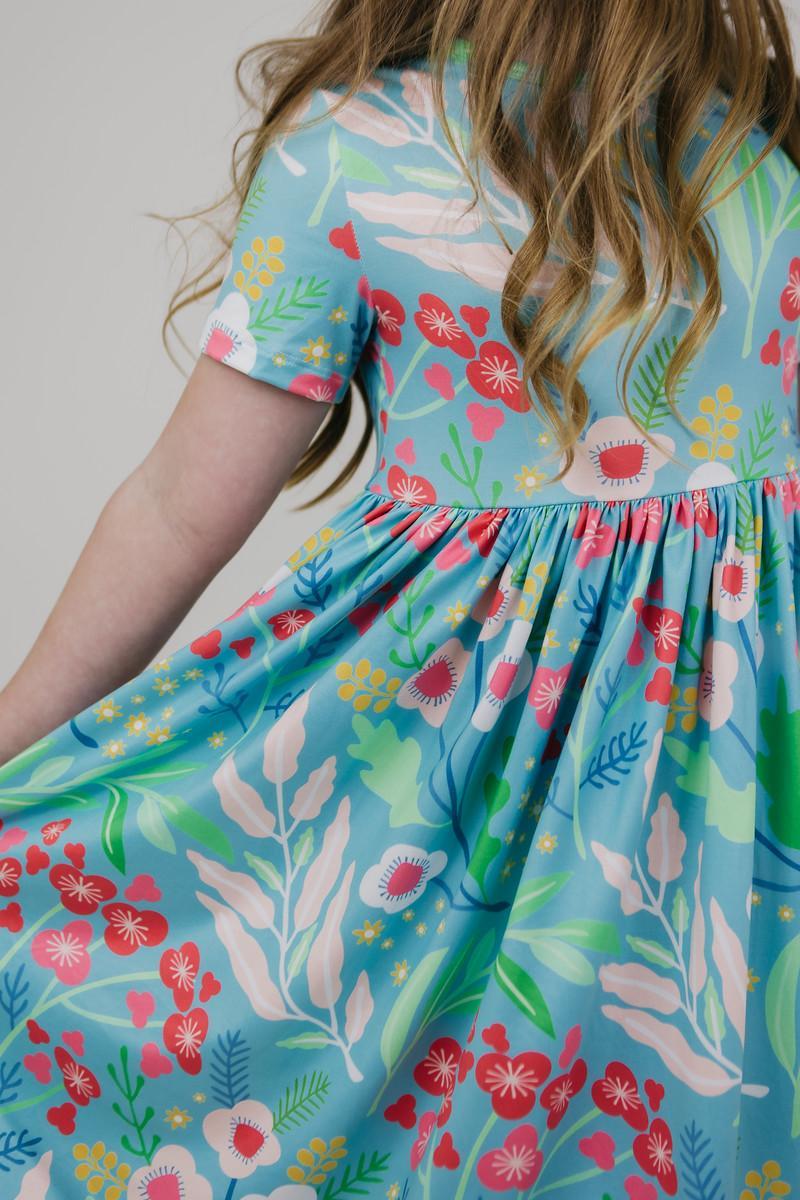 A girl twirls in the Spring Breeze S/S Pocket Twirl Dress with a focus on the back, emphasizing the colorful floral print and twirling skirt.