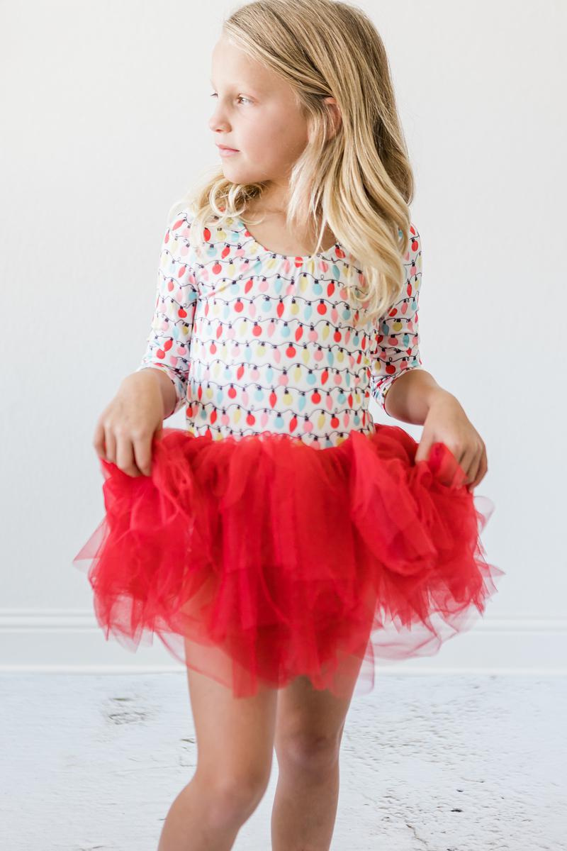 A girl standing gracefully in a Merry & Bright 3/4 Tutu Leotard for Girls, showcasing the fluffy red tutu and festive light bulb pattern.