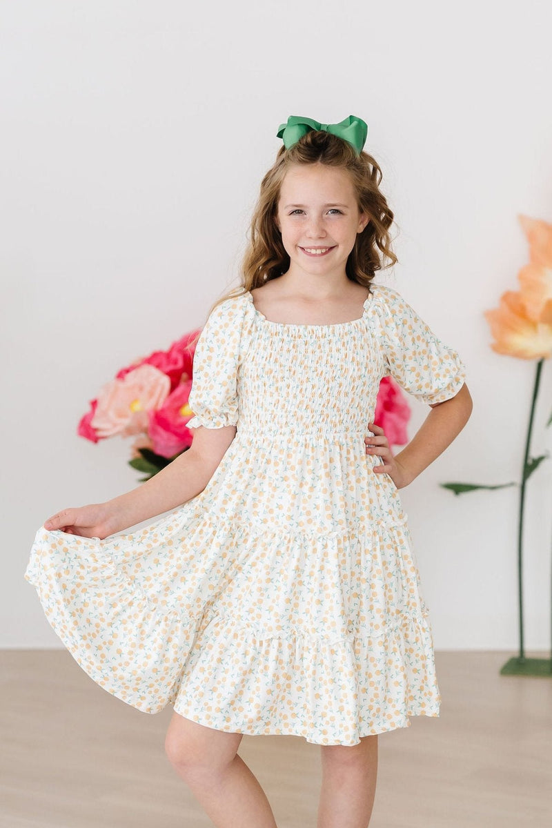 A smiling girl wearing the Marvelous Marigolds Smocked Ruffle Dress by Mila & Rose, holding her skirt and standing against a colorful floral setting.