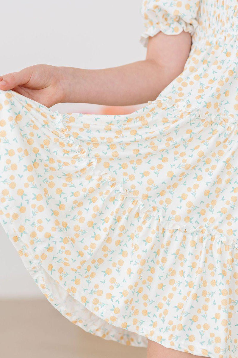 Close-up of the Marvelous Marigolds Smocked Ruffle Dress featuring a delicate floral print with yellow marigold flowers on a soft white background, highlighting the playful ruffle skirt and smocked bodice.