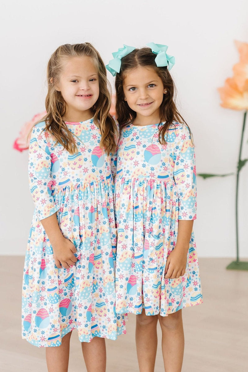 Two cheerful girls pose together in their matching Egg-cellent 3/4 Sleeve Pocket Twirl Dresses, radiating fun and friendship as they celebrate their style.