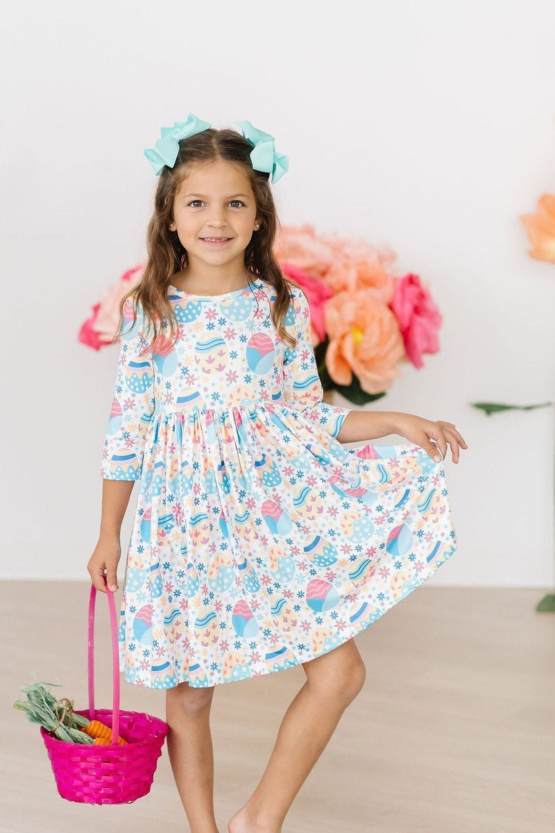 A little girl holds a colorful basket and poses in her Egg-cellent 3/4 Sleeve Pocket Twirl Dress, embodying a charming and whimsical atmosphere.