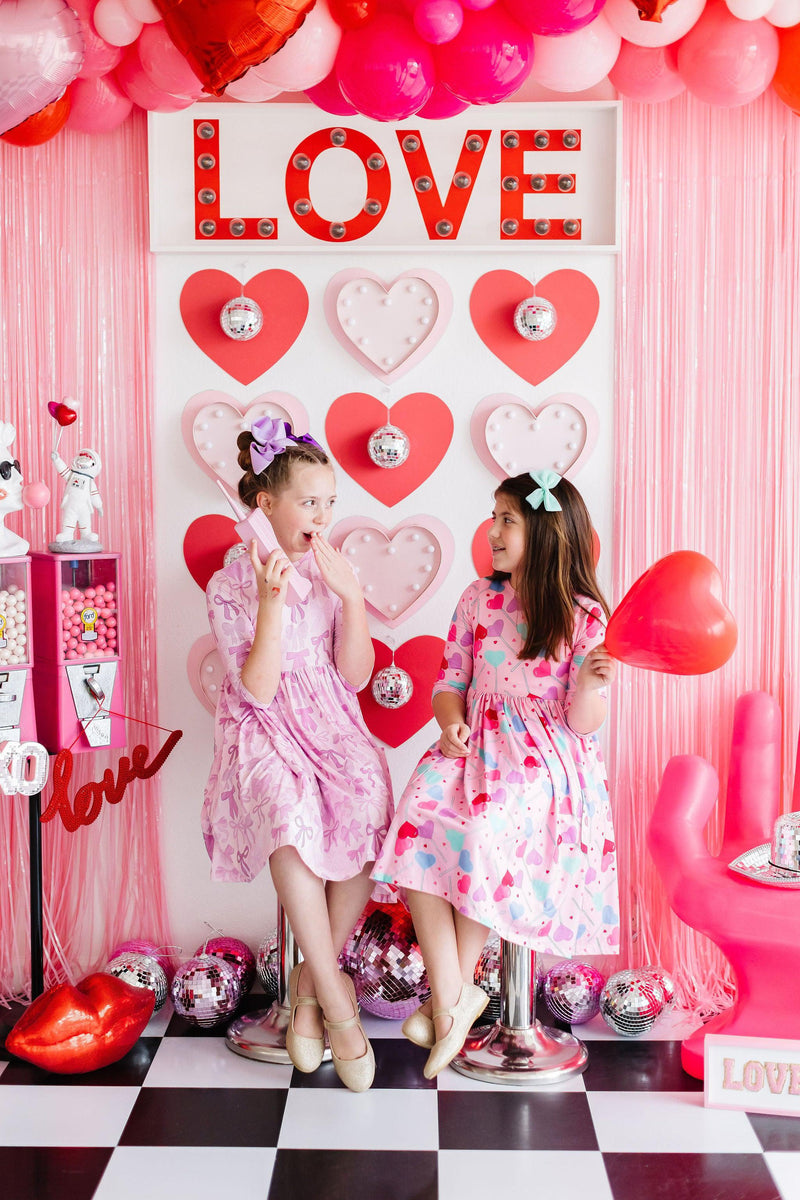 Twinkle Ties 3/4 Sleeve Pocket Twirl Dress worn by two girls enjoying a fun moment together, surrounded by heart-themed decor, highlighting the dress's stylish appearance and suitability for playful occasions.