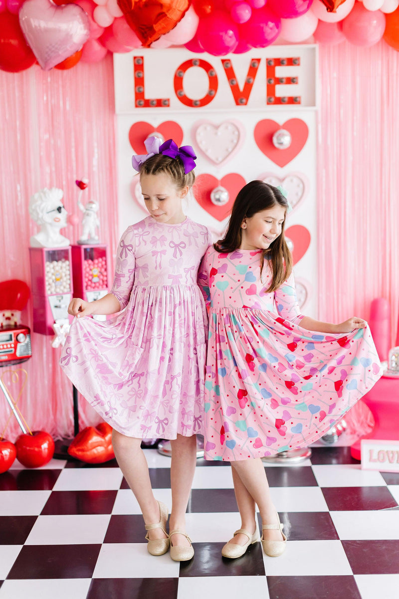 Two girls happily twirling in their Twinkle Ties 3/4 Sleeve Pocket Twirl Dresses from Mila & Rose, surrounded by festive decorations in shades of pink.