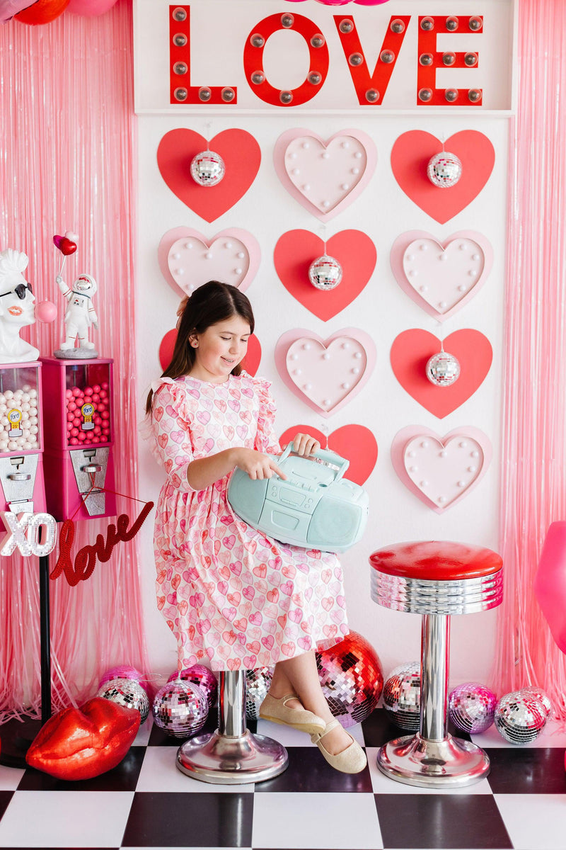 A young girl in the Melt My Heart 3/4 Sleeve Ruffle Twirl Dress, enjoying a fun moment with a cute teal boombox. She sits beside vibrant heart decorations, showcasing the dress's charming design that invites smiles and twirls at any playful gathering!