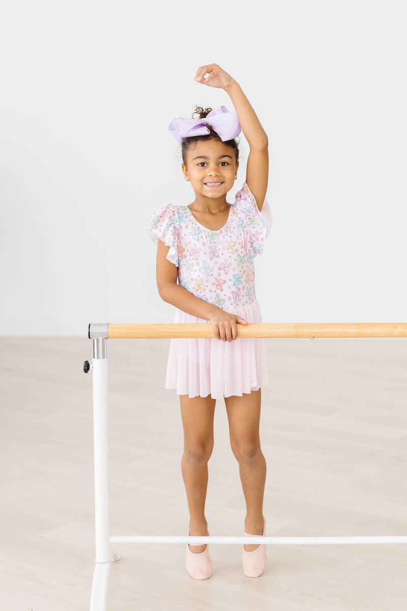 A child posing in the Springtime Bows Short Sleeve Twirl Leotard by Mila & Rose, demonstrating a ballet pose at the barre with a cheerful expression.