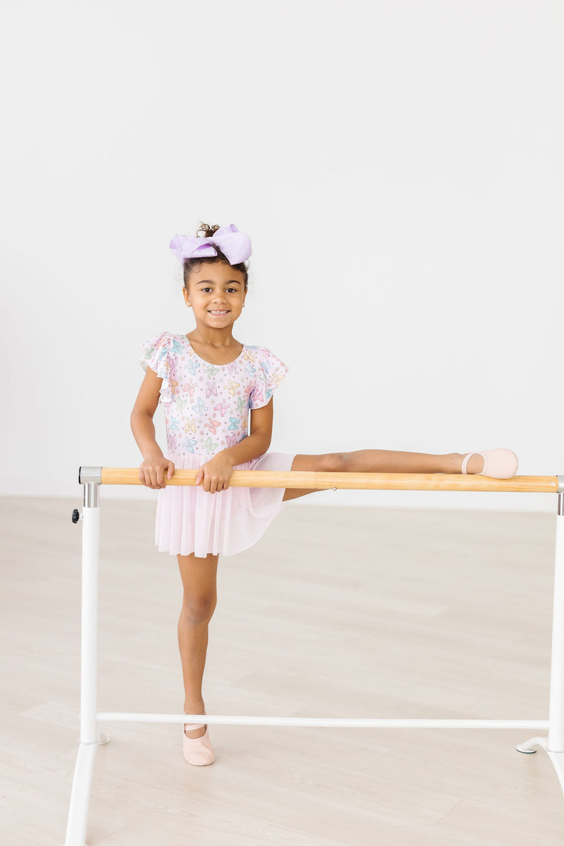 A young girl performing an elegant ballet pose in the Springtime Bows Short Sleeve Twirl Leotard by Mila & Rose, complete with pink flats and a big bow in her hair.