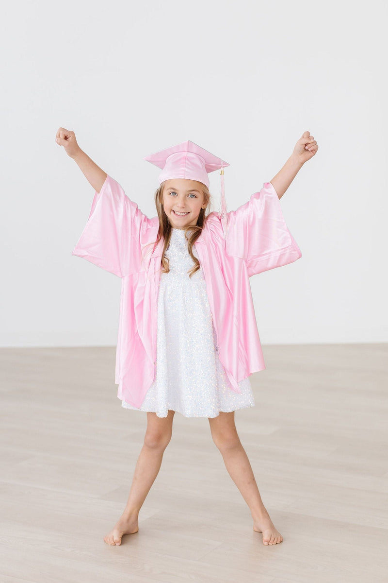 A girl celebrating in a pink graduation cap and gown over a white sequin party dress, embodying joy and achievement.