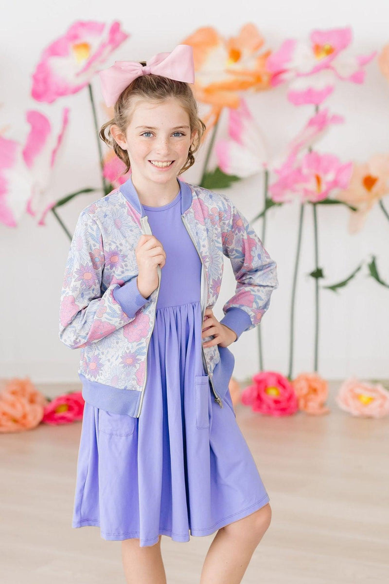 Stylish child wearing the Periwinkle S/S Pocket Twirl Dress paired with a floral bomber jacket, posing joyfully among vibrant flower decorations.