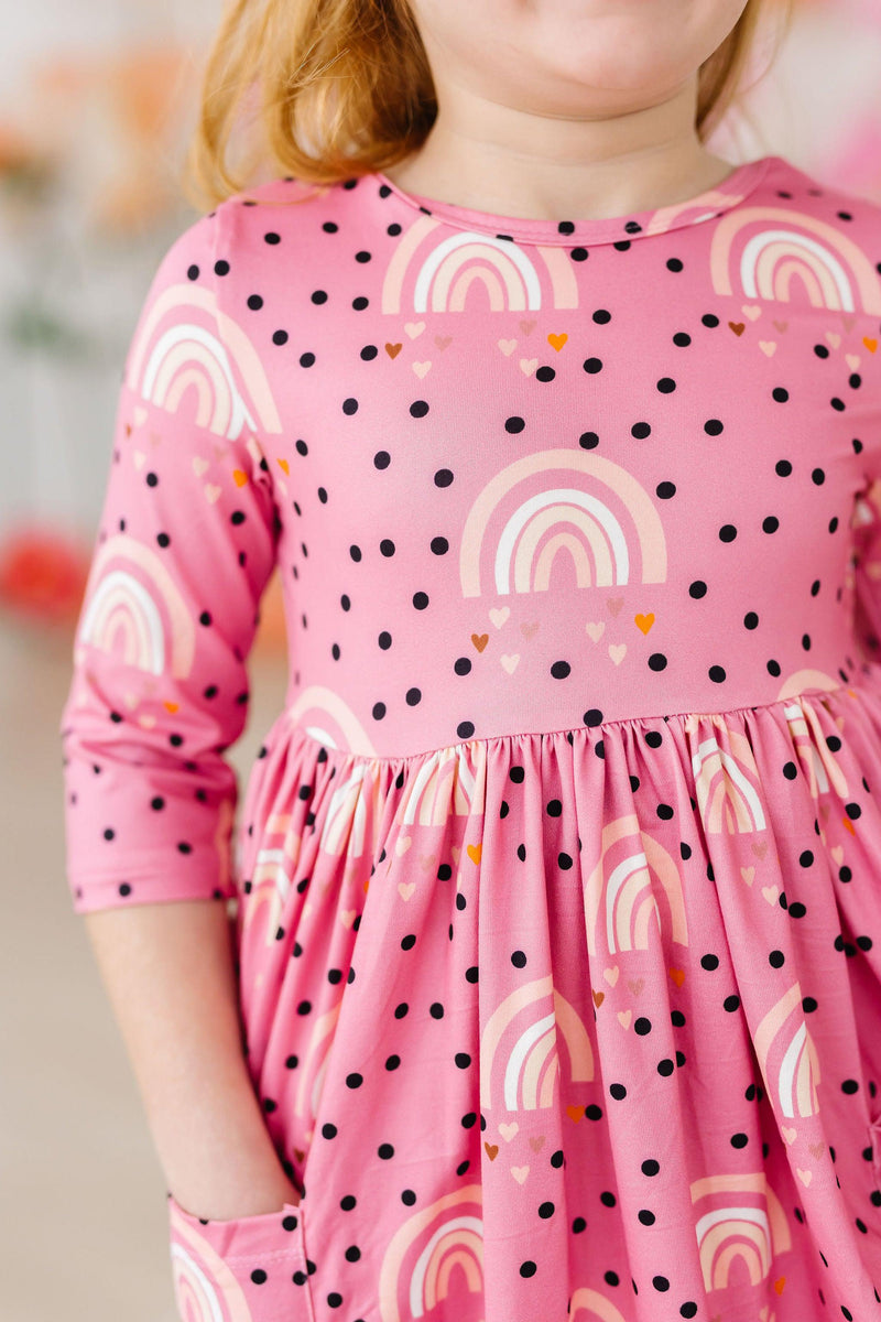 Girl wearing the Rainbow Wishes 3/4 Sleeve Pocket Twirl Dress by Mila & Rose®, showcasing a playful pink design with rainbows and polka dots.