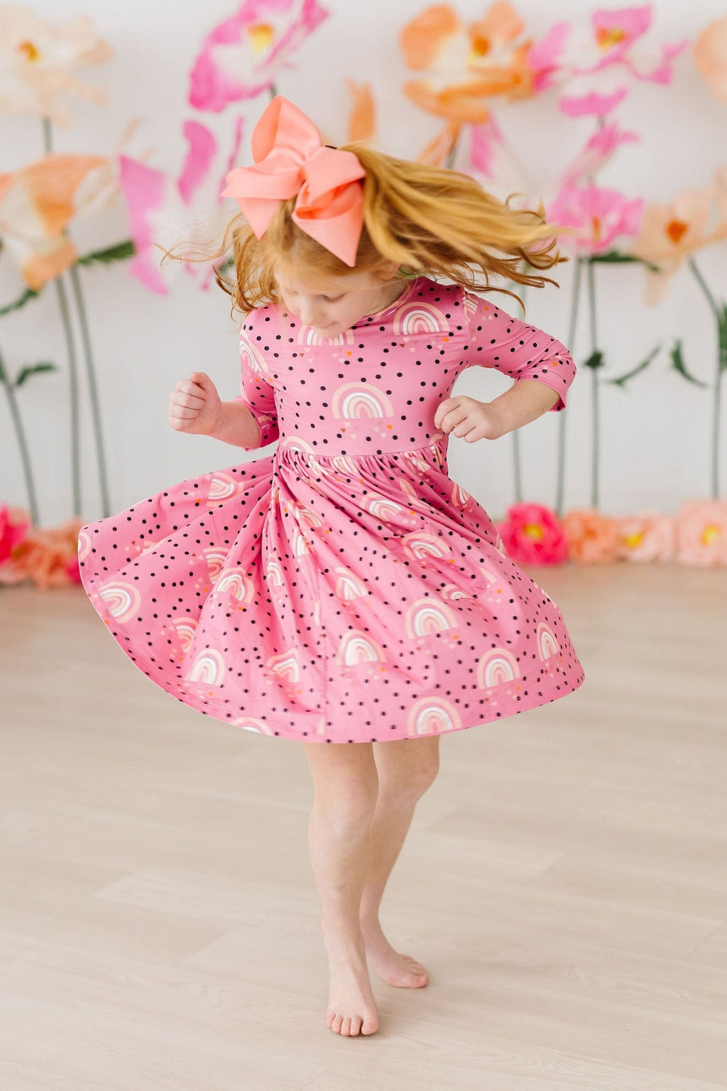 A young girl wearing the Rainbow Wishes 3/4 Sleeve Pocket Twirl Dress by Mila & Rose, twirling joyfully in a pink dress adorned with rainbow and polka dot patterns, perfect for playful occasions.