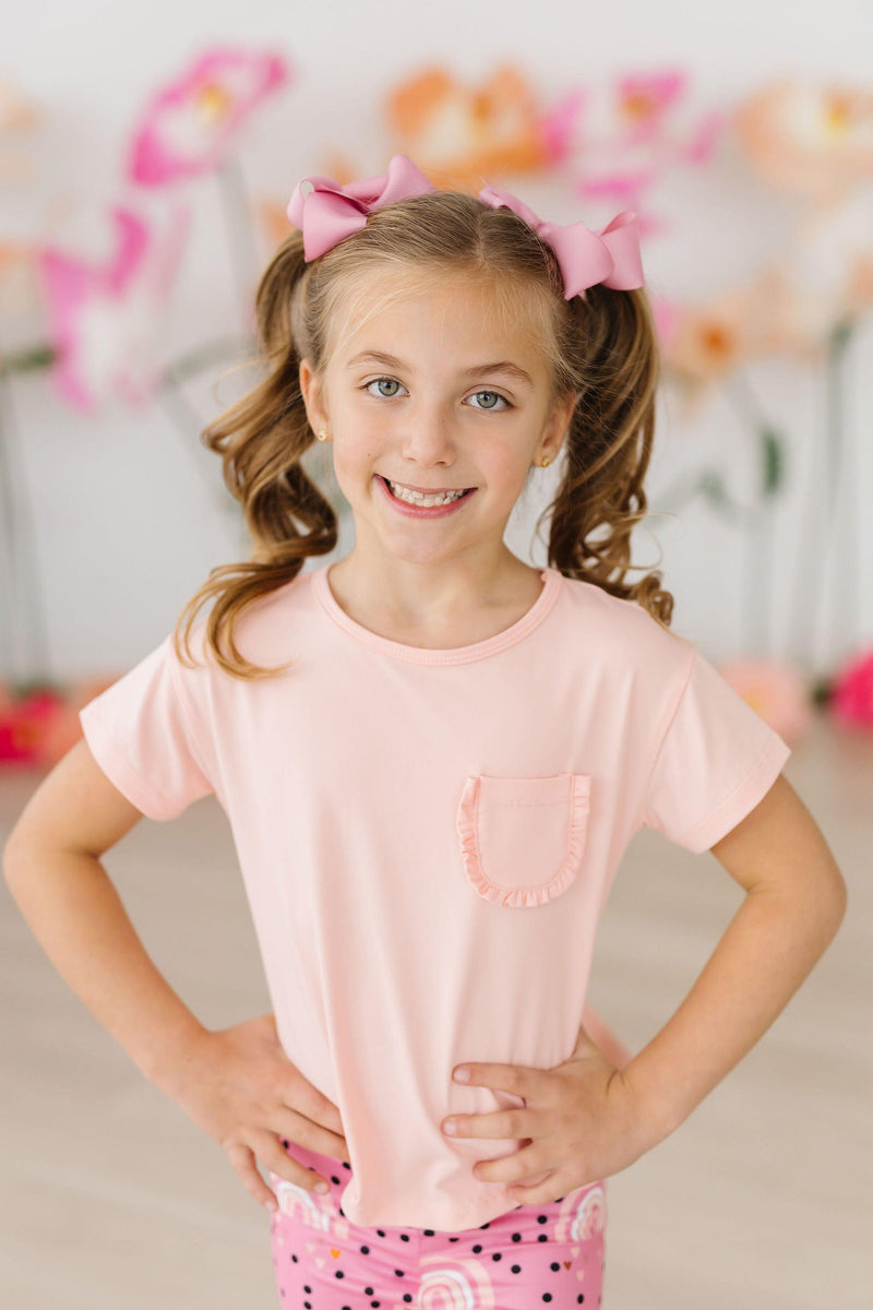 Smiling girl with pigtails wearing the Peach Ruffle Pocket Tee, radiating joy and charm in a vibrant setting.