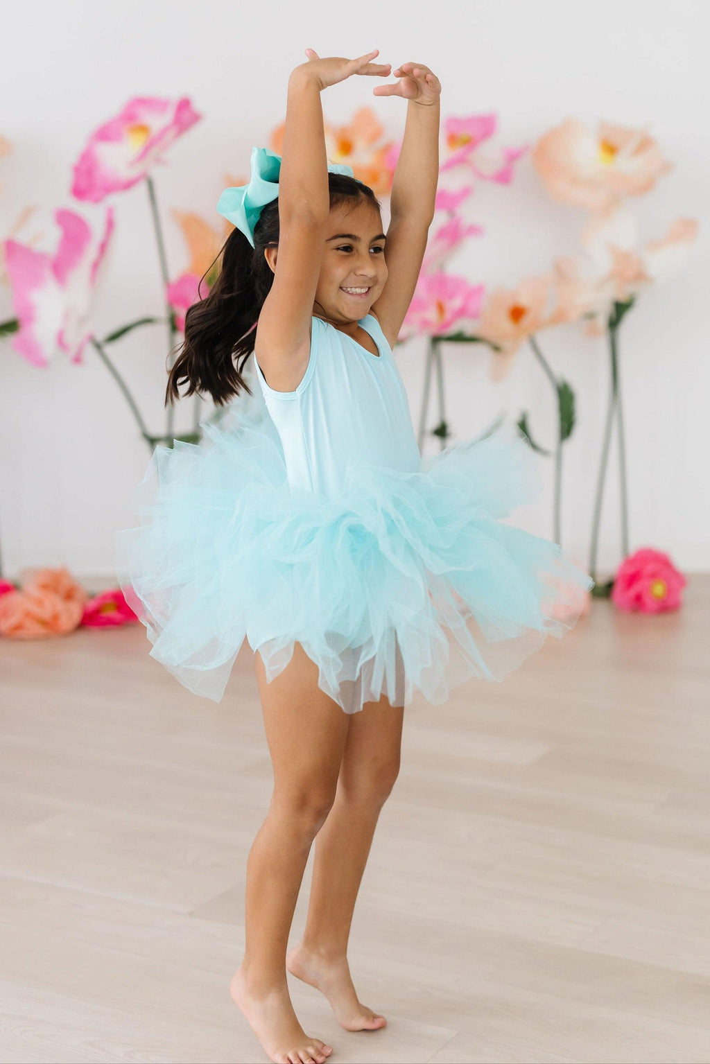 A young girl joyfully dancing in an Aqua Tank Tutu Leotard, featuring a fluffy aqua tutu, set against a backdrop of colorful floral decorations.