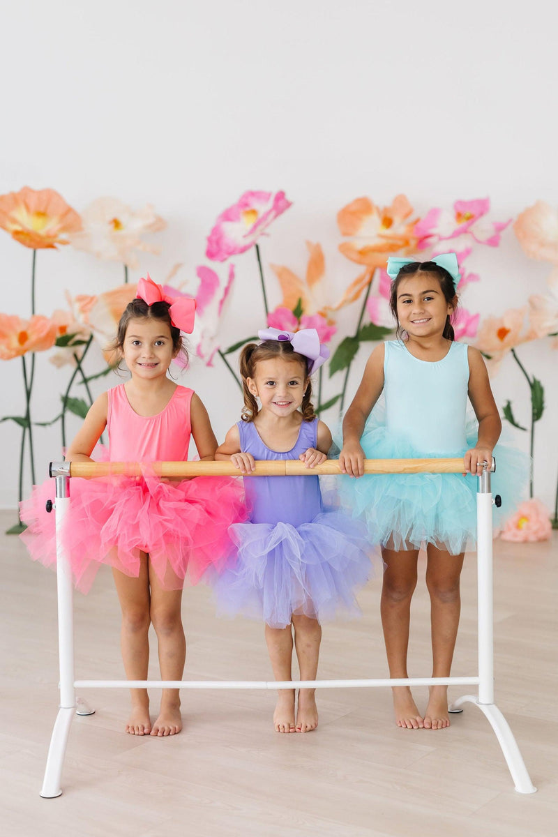 Three girls wearing colorful Aqua Tank Tutu Leotards in pink, purple, and aqua, standing at a ballet barre with a backdrop of oversized flowers.