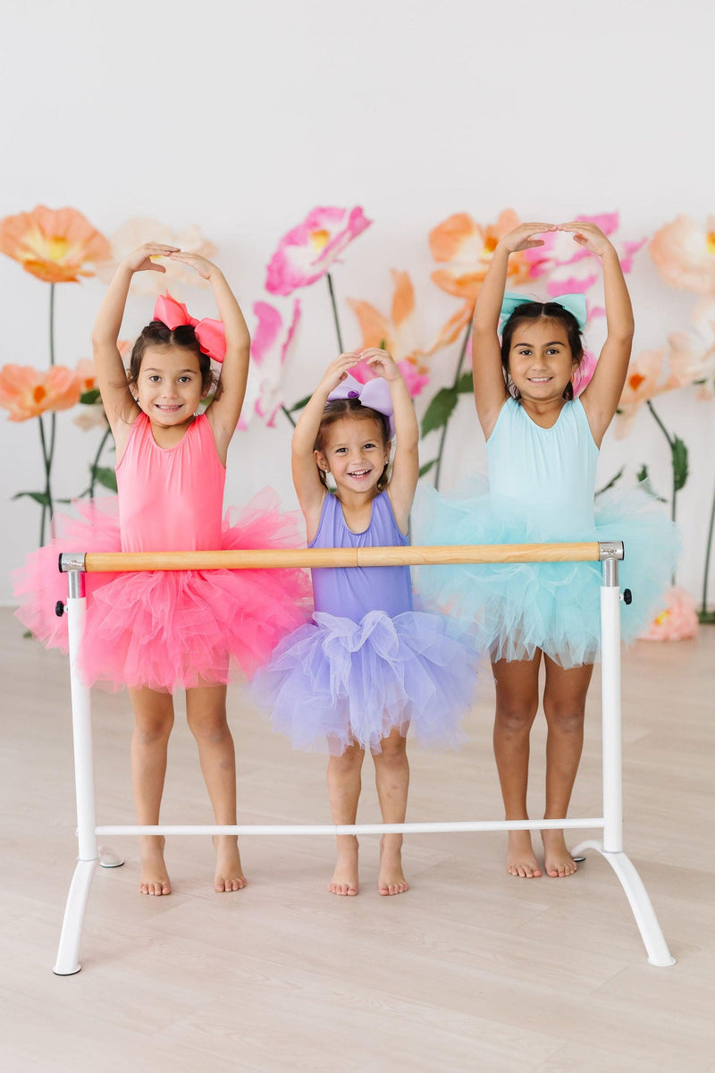Three girls showcasing their Aqua Tank Tutu Leotards in pink, purple, and aqua, joyfully posing together at a ballet barre surrounded by flower decorations.