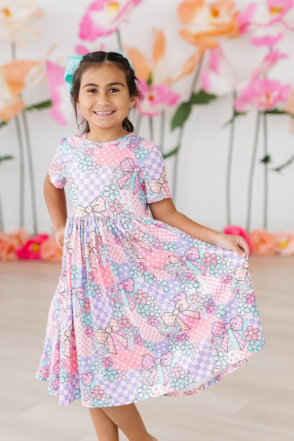 Happy little girl spinning in the Patchwork Princess S/S Pocket Twirl Dress by Mila & Rose, surrounded by colorful floral backdrop.