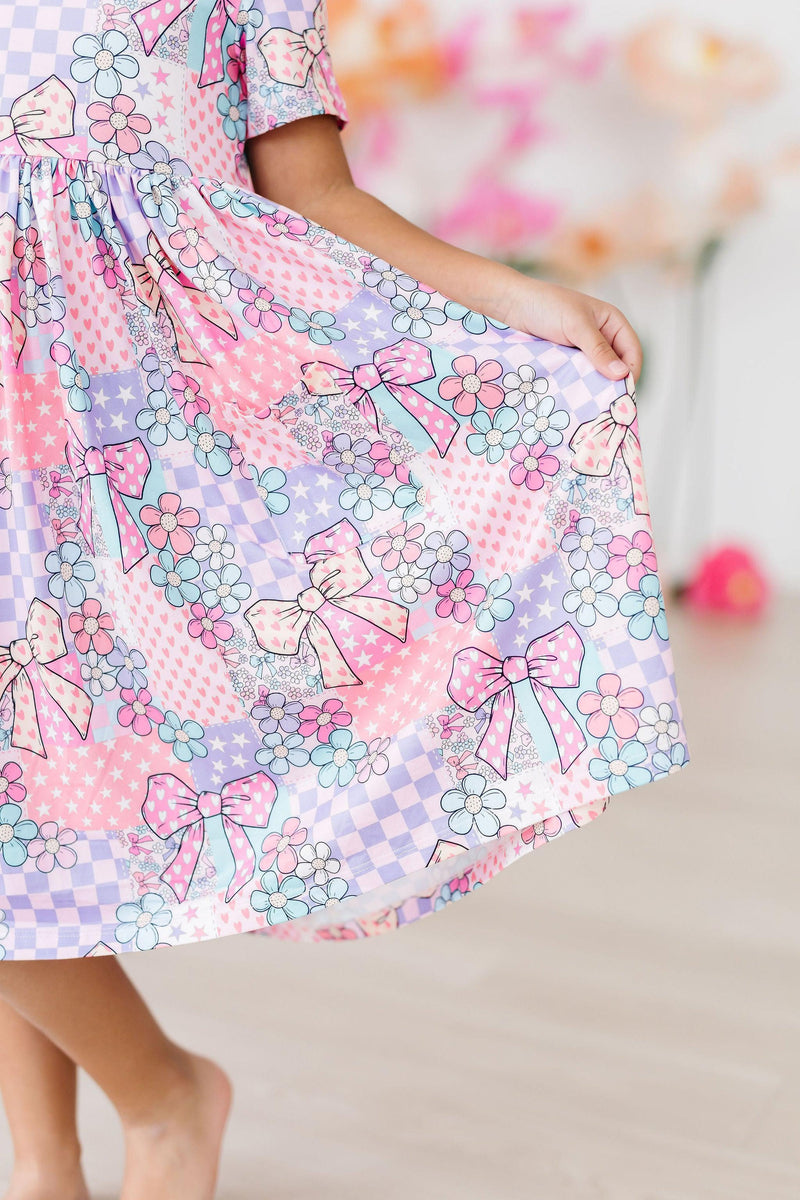 A close-up look at the enchanting Patchwork Princess S/S Pocket Twirl Dress featuring colorful floral patterns and bows, perfect for twirling and fun adventures.