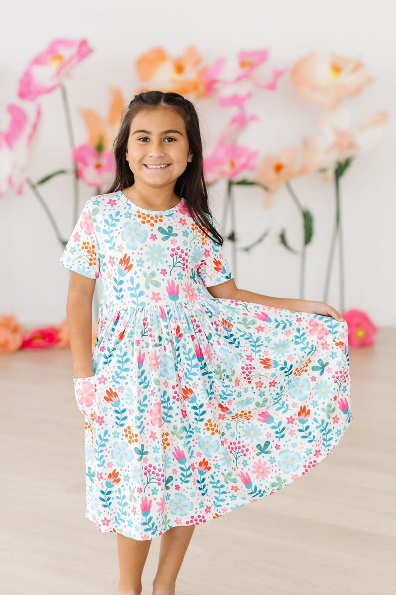 A smiling girl in the Sunlit Garden S/S Pocket Twirl Dress with pockets, standing happily amidst vibrant flower decorations.
