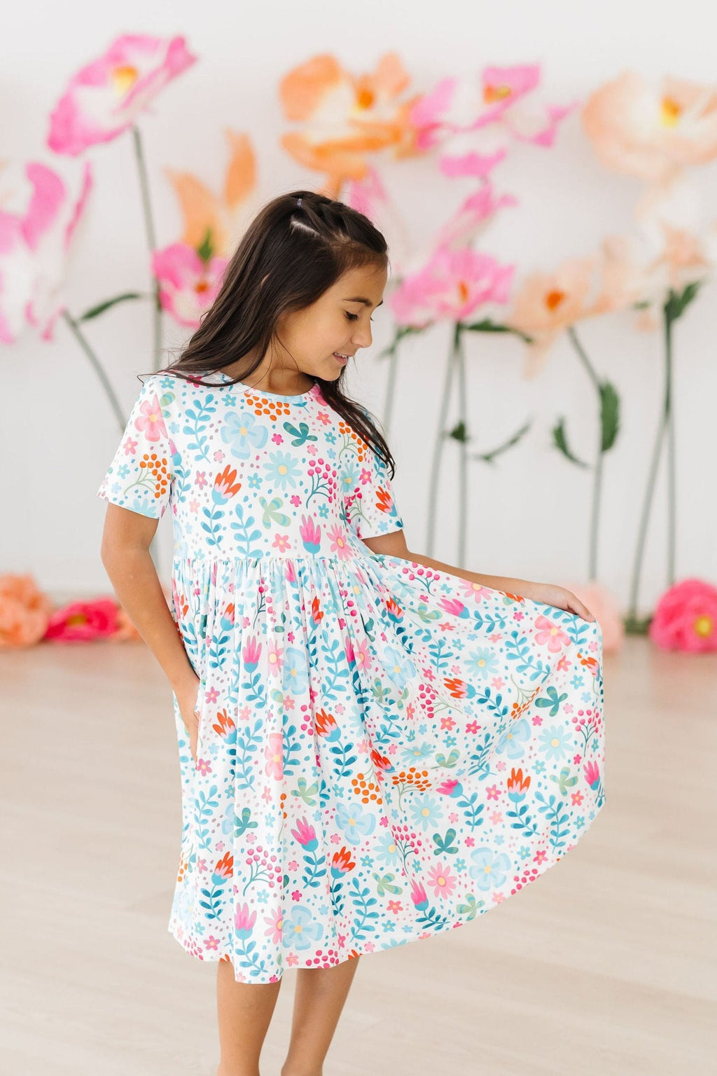 A young girl joyfully twirling in the Sunlit Garden S/S Pocket Twirl Dress, featuring vibrant floral patterns, perfect for playtime and special events.