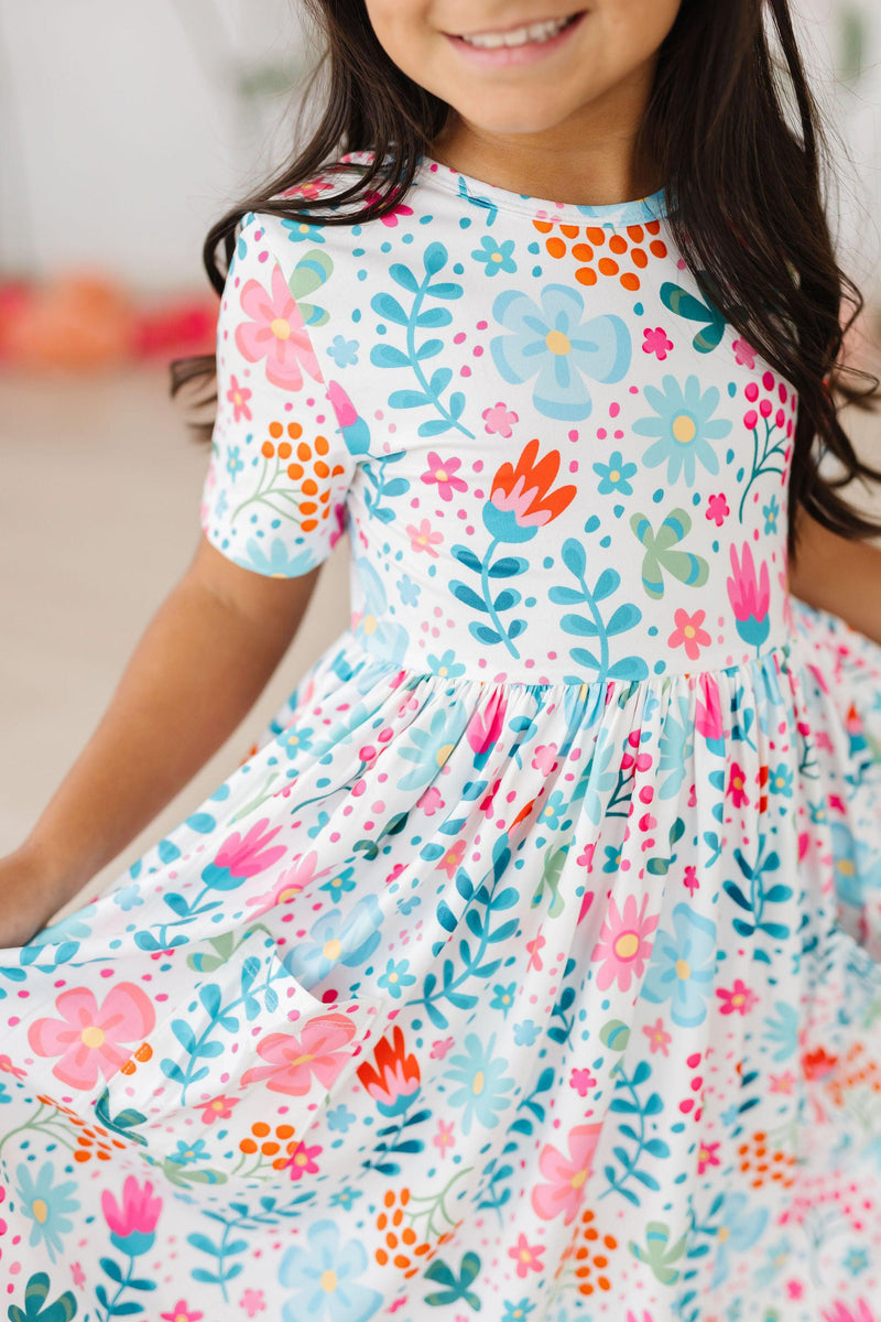 A joyful girl wearing the Sunlit Garden S/S Pocket Twirl Dress, showcasing a colorful floral print and twirling in delight.