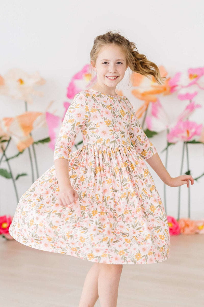 Adorable girl twirling in the Pretty Peachy 3/4 Sleeve Pocket Twirl Dress, surrounded by colorful flowers.