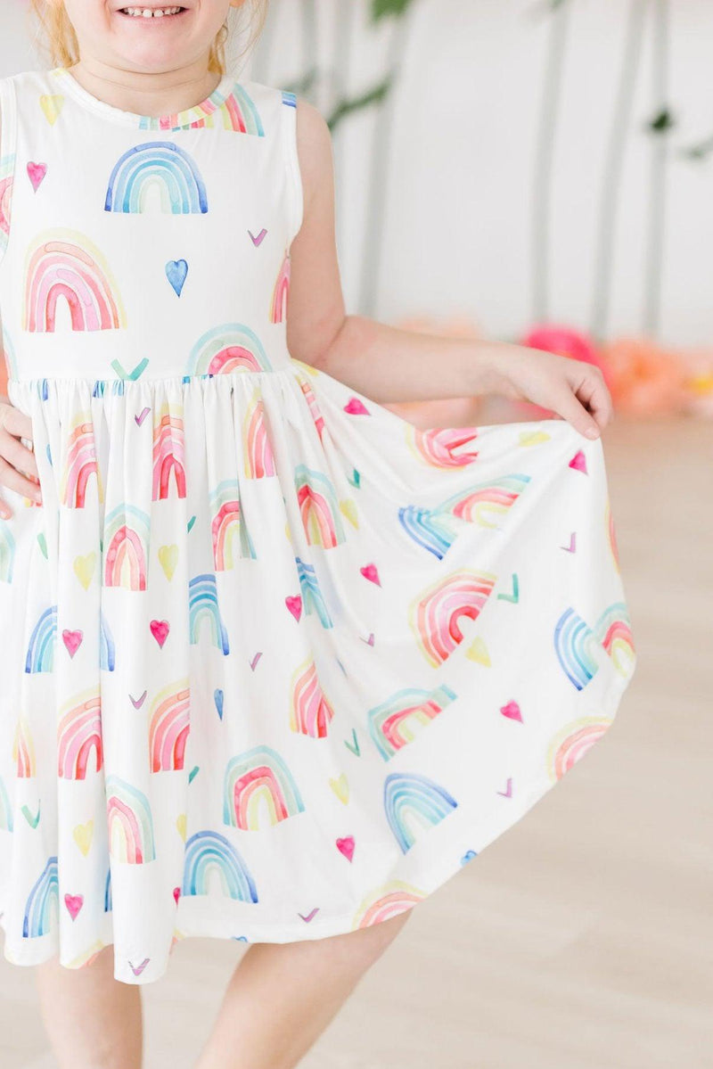 A young girl wearing the Radiant Rainbows Tank Twirl Dress, joyfully lifting the skirt while smiling, showcasing the colorful rainbow and heart patterns.