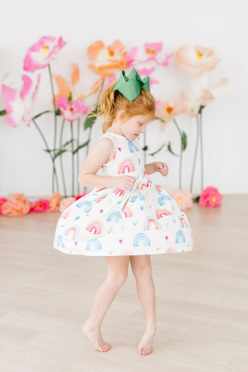 A young girl spinning playfully in the Radiant Rainbows Tank Twirl Dress, demonstrating its twirl design and bright colors against a backdrop of faux flowers.