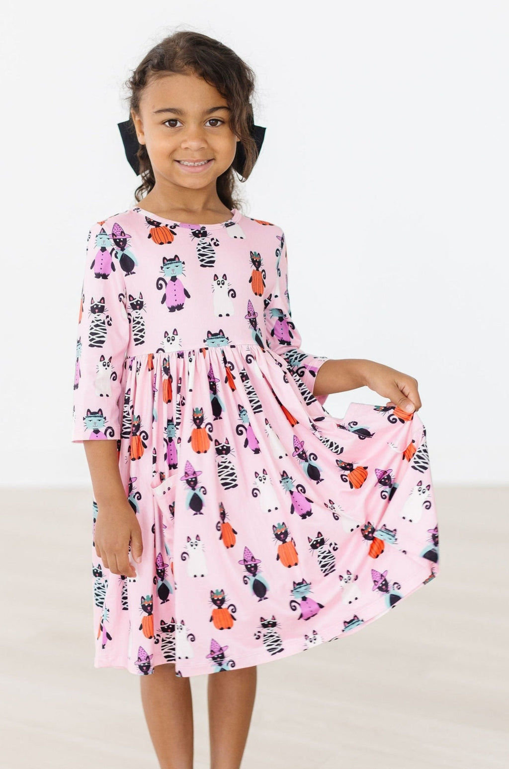 A happy young girl wearing the Purr-fectly Spooky 3/4 Sleeve Pocket Twirl Dress, featuring playful Halloween-themed designs. She is joyfully twirling, showcasing the dress's elegant flow and vibrant colors.