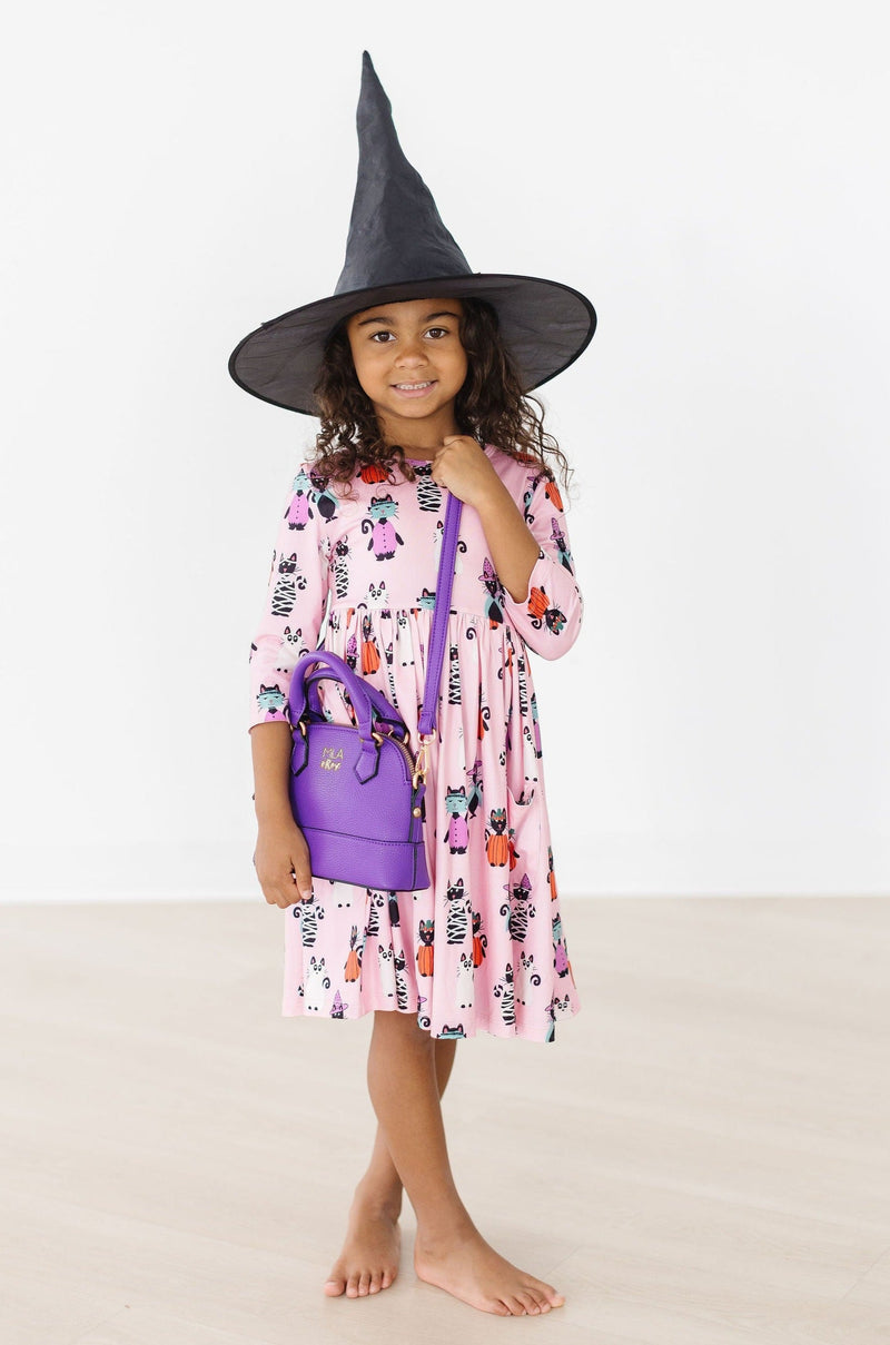 A charming young girl dressed in the Purr-fectly Spooky 3/4 Sleeve Pocket Twirl Dress, complemented by a witch hat and purple handbag, embodying a fun Halloween spirit.