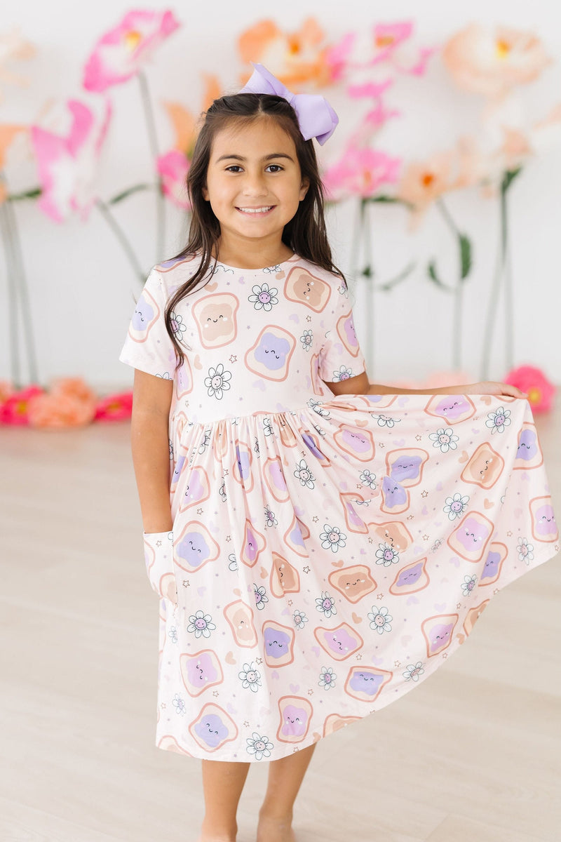 A smiling girl twirling in her Peanut Butter Jelly Time S/S Pocket Twirl Dress by Mila & Rose, showcasing the charming peanut butter and jelly print in a beautifully bright floral setting.