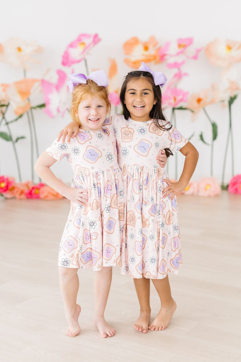 Two adorable friends posing confidently in their Peanut Butter Jelly Time S/S Pocket Twirl Dresses by Mila & Rose, surrounded by vibrant flowers that perfectly match their cheerful outfits.